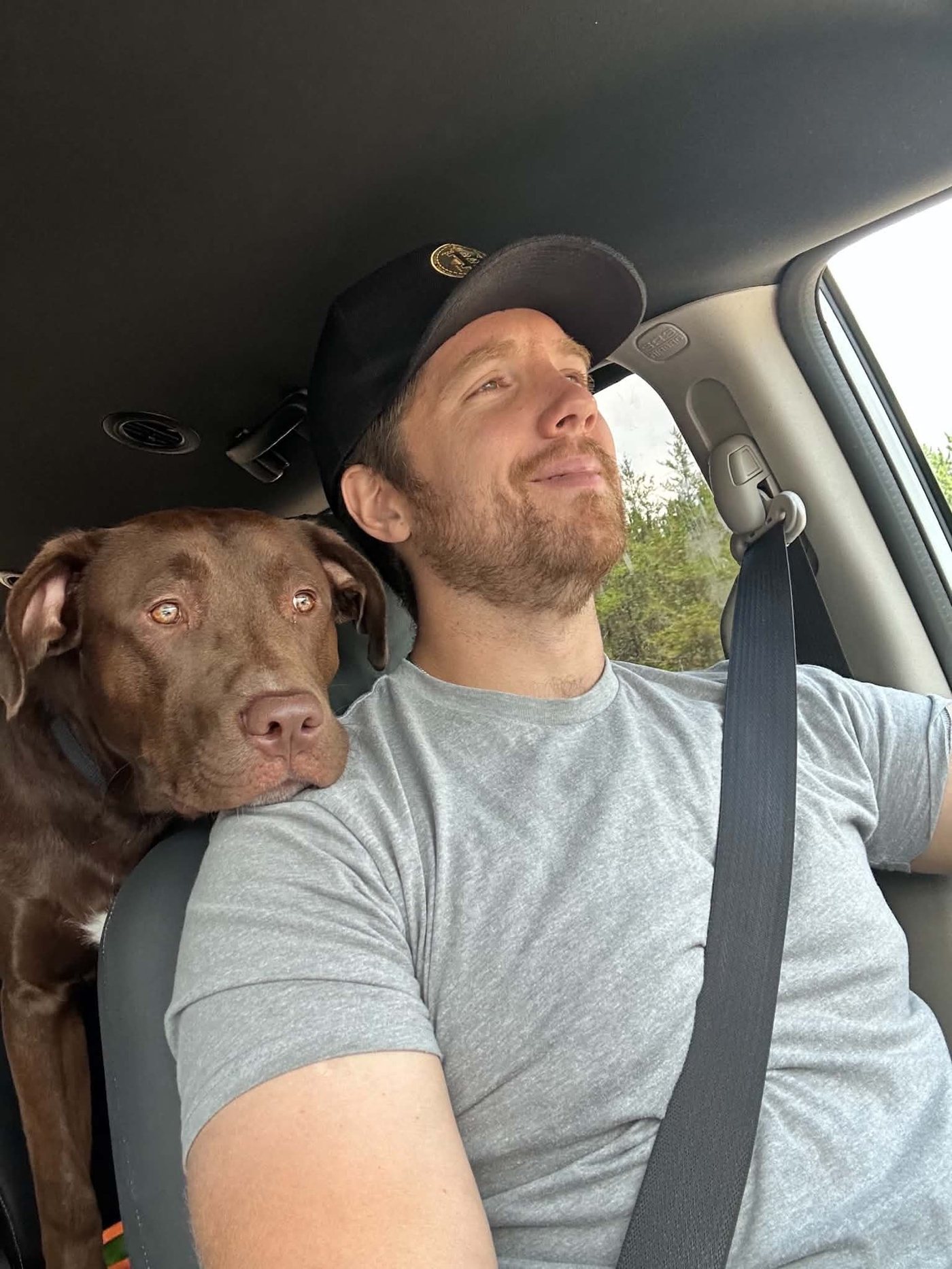 Elliott in the passenger seat, with a brown dog resting its head on his shoulder.